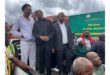 A group of people stand closely together, smiling and chatting. They're outdoors near a sign for Moutse Traffic Station. The mood is friendly.