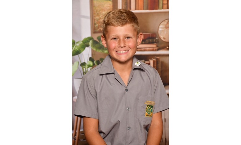 A boy with short, light brown hair, smiles in a school uniform featuring a gray shirt with a yellow-green crest. Background shows a cozy room with plants and books.