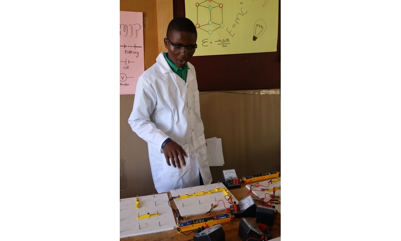 A student in a lab coat explains a science project involving circuits and batteries on a table. Charts and diagrams are displayed on the wall.