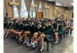 A group of students in green uniforms sit on chairs in a classroom, wearing virtual reality headsets. A person stands at the back, guiding them.