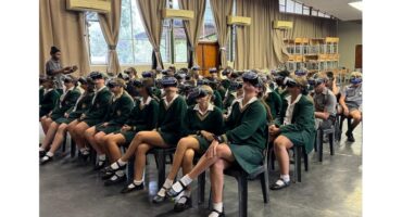 A group of students in green uniforms sit on chairs in a classroom, wearing virtual reality headsets. A person stands at the back, guiding them.