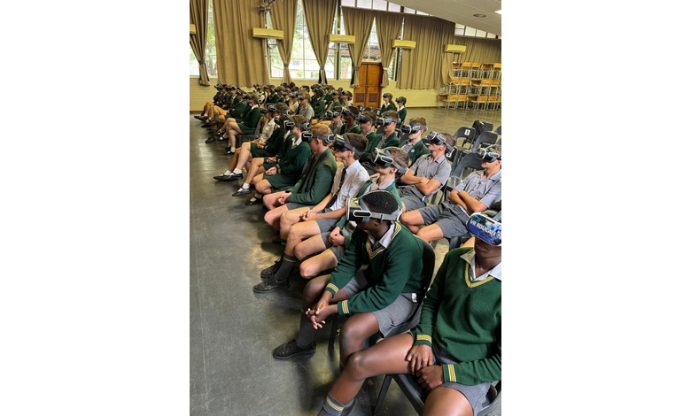 Students in a school hall wearing virtual reality headsets, seated in rows. They're in uniform, surrounded by beige curtains, with a focused atmosphere.