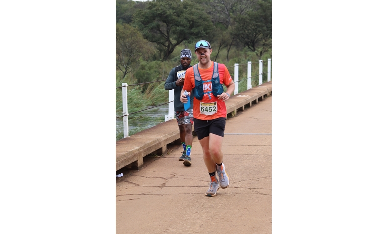 A man in an orange shirt and black shorts runs on a paved path over a bridge, smiling and holding water bottles. Another runner follows behind. The scene is set outdoors with lush greenery and a calm, determined atmosphere.