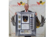 A man in a suit holding a framed police memorabilia collection featuring badges, a cap, medals, and a certificate. "Hawks Directorate" banners in the background.