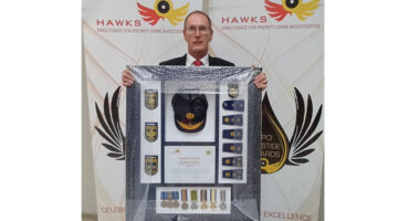 A man in a suit holding a framed police memorabilia collection featuring badges, a cap, medals, and a certificate. "Hawks Directorate" banners in the background.