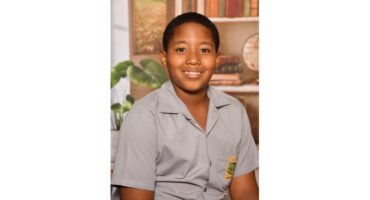 Smiling boy in a school uniform sits in front of a backdrop featuring books and plants, conveying a sense of warmth and education.