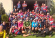 A large group of cyclists in colorful jerseys and helmets pose together outside a building on a sunny day, exuding camaraderie and enthusiasm.