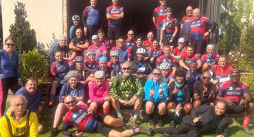 A large group of cyclists in colorful jerseys and helmets pose together outside a building on a sunny day, exuding camaraderie and enthusiasm.