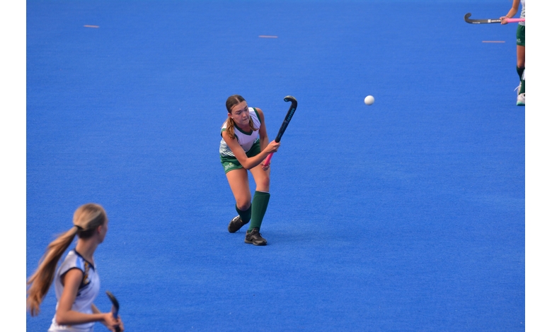 A female field hockey player in a green uniform strikes a ball with focus on a vibrant blue turf, with teammates in the background. The scene conveys intensity and athleticism.