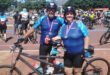 A smiling man and woman in matching blue cycling gear stand with their tandem bike, medals around their necks, amid a lively crowd of cyclists.
