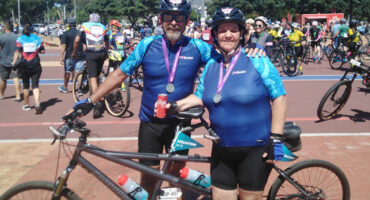 A smiling man and woman in matching blue cycling gear stand with their tandem bike, medals around their necks, amid a lively crowd of cyclists.