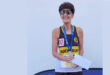 An older woman wearing sunglasses and a running bib proudly holds a certificate. She has a medal, smiling against a plain background, conveying joy and achievement.
