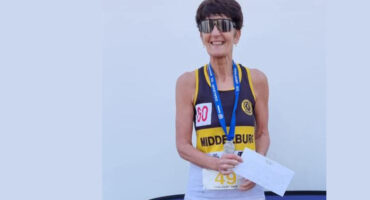 An older woman wearing sunglasses and a running bib proudly holds a certificate. She has a medal, smiling against a plain background, conveying joy and achievement.