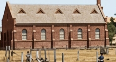 A historic, red-brick building with arched windows and a gabled roof stands in a dry, grassy area. Two people in period clothing are in the foreground.