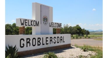 Welcome sign for Groblersdal against a clear blue sky. The sign features "Welkom" and "Welcome" with greenery and a road in the background.
