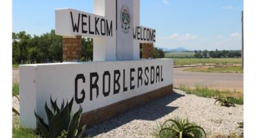 A roadside sign reads “Welkom Welcome Groblersdal” with a crest above. It’s set against a rural landscape, blue sky, and greenery, evoking a welcoming tone.