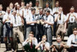 A group of young men in vintage attire gather on stairs, enjoying drinks and snacks. They wear suspenders and ties, exuding a joyful, camaraderie-filled atmosphere.
