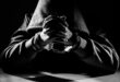 A hooded figure in handcuffs sits at a table, hands clasped. The black-and-white image evokes a tense, mysterious atmosphere with dramatic lighting.