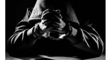A hooded figure in handcuffs sits at a table, hands clasped. The black-and-white image evokes a tense, mysterious atmosphere with dramatic lighting.