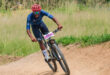 Cyclist in a red helmet and blue outfit races on a dirt trail, exuding determination. Number 704 is visible on the bike. Green grass in the background.