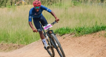 Cyclist in a red helmet and blue outfit races on a dirt trail, exuding determination. Number 704 is visible on the bike. Green grass in the background.