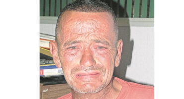 A man with short hair and a weathered face appears emotional, with furrowed brows and teary eyes. He wears a red shirt, sitting indoors near books.