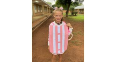 Child dressed as a pop-tart with cat ears, whiskers, and a joyful expression. The background shows a schoolyard and a path lined with trees.