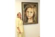 A woman in a yellow dress stands smiling beside a large, framed portrait of a young girl with long hair. The portrait conveys a serious tone.
