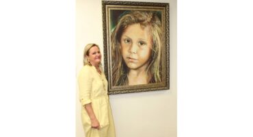 A woman in a yellow dress stands smiling beside a large, framed portrait of a young girl with long hair. The portrait conveys a serious tone.