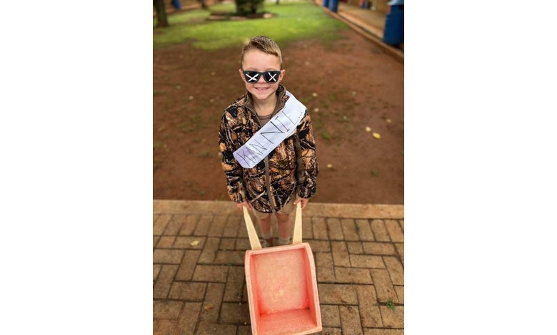 A smiling child in camo clothing, wearing cross-shaped sunglasses and a sash, pulls an empty wheelbarrow on a brick path. The scene feels playful.