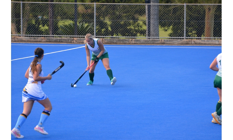 A field hockey match on a blue turf with players wearing green and white, and white and blue uniforms. One player is in mid-action, focusing on the ball.