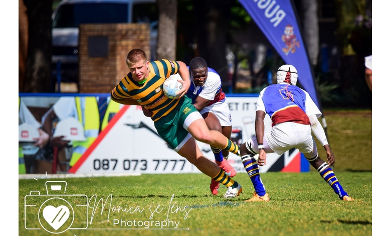 Rugby player in green and yellow stripes sprints forward, clutching the ball. Opponent in blue and red attempts a tackle. Intense action on grassy field.