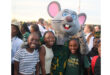 A large mouse mascot with big pink ears and blue eyes poses with smiling children at an outdoor event. The mood is joyful and festive.