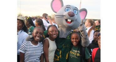 A large mouse mascot with big pink ears and blue eyes poses with smiling children at an outdoor event. The mood is joyful and festive.
