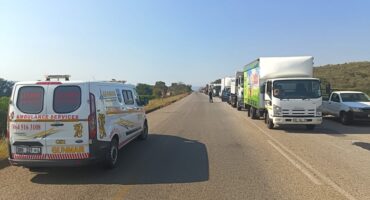 A long line of trucks and an ambulance on a clear highway under a bright sky. The vehicles are heading in the same direction, suggesting a traffic delay.