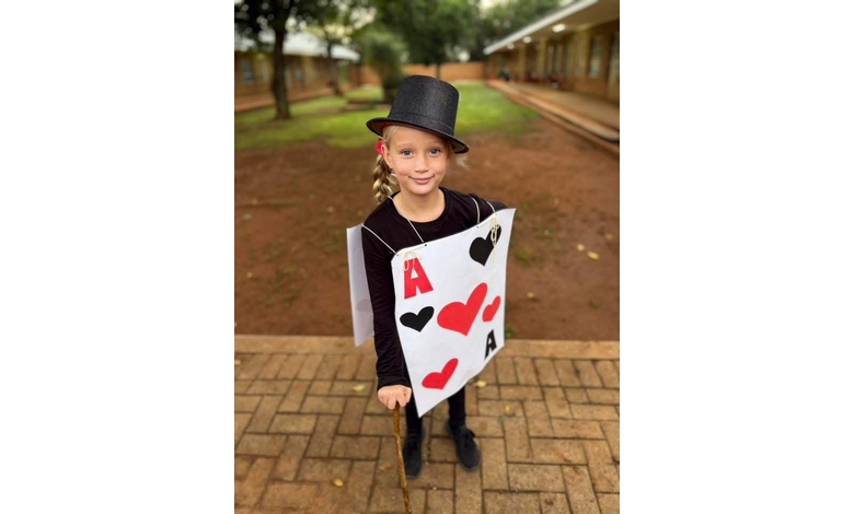 A young child dressed as a playing card with hearts and spades, wearing a black top hat, holds a cane, smiling outdoors on a brick pathway.