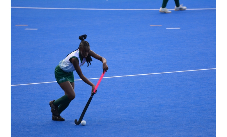 A field hockey player in a green and white uniform skillfully maneuvers the ball on a bright blue turf, focused and in action, conveying intensity.