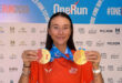 Smiling woman in an orange "One Run" shirt holds two medals against a backdrop with event logos. The atmosphere is celebratory and triumphant.