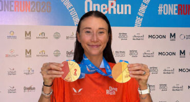 Smiling woman in an orange "One Run" shirt holds two medals against a backdrop with event logos. The atmosphere is celebratory and triumphant.
