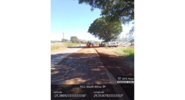 Road construction on N11, South Africa. Workers in orange uniforms operate machinery under a large tree. Clear blue sky, time and GPS data overlay.