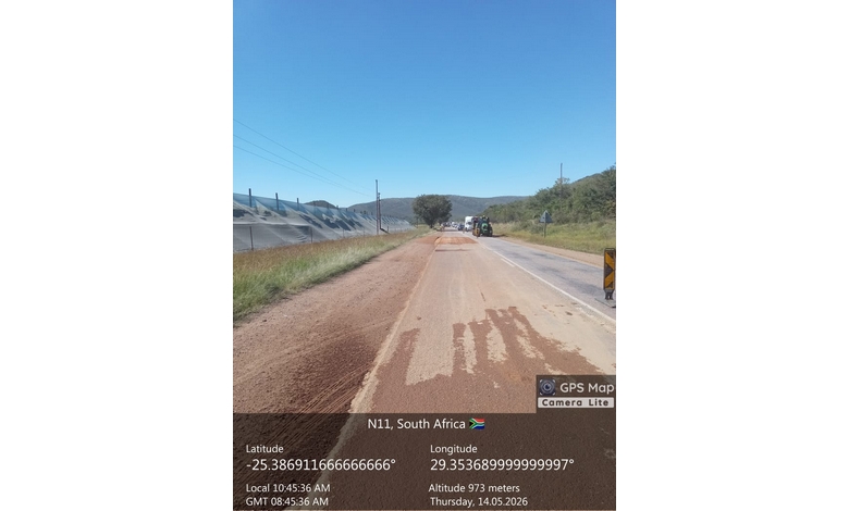 Roadwork on N11, South Africa, under a clear blue sky. A road crew works on a dirt patch. Surrounding are green fields and distant hills.