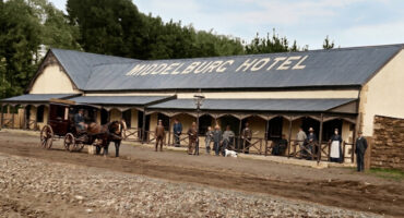 A vintage scene of Middelburg Hotel with dirt road. People in period clothing stand outside, alongside a horse-drawn carriage, evoking a historical atmosphere.