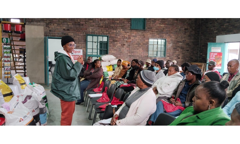 A person in a green jacket and beanie speaks to a seated audience in a brick-walled room. The attendees, warmly dressed, listen attentively. Bags of feed are stacked at the side.