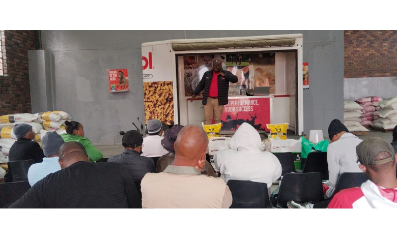 A person in a red jacket speaks to an engaged audience seated in a warehouse setting. Stacks of feed bags are visible. The mood is informative.