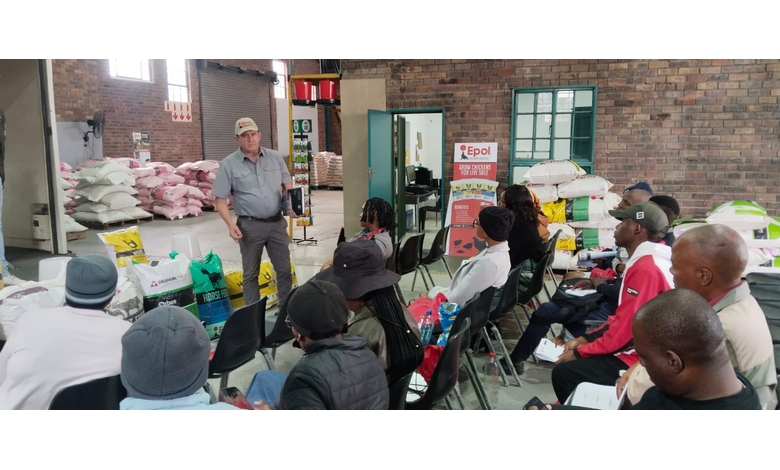 A man in a cap and uniform addresses a seated group indoors. Bags of feed are stacked on the left. The atmosphere is attentive and educational.