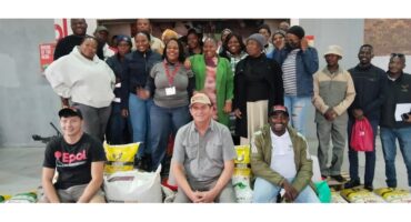 A diverse group of people smiling, gathered indoors. They stand and sit around stacks of feed bags, conveying a sense of community and teamwork.