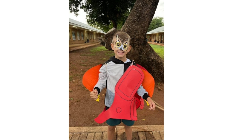A child stands outdoors wearing an owl mask, cape, and a red and orange rocket prop. They smile, surrounded by trees and school buildings. Playful mood.