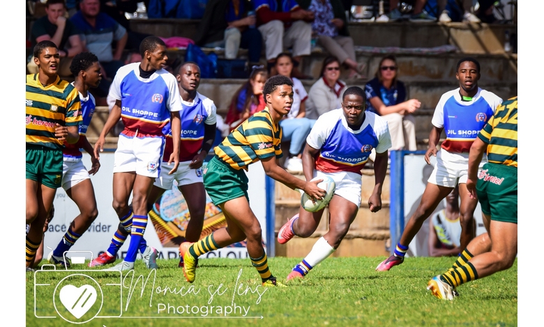 Rugby match action with a player in a green and yellow striped jersey running with the ball, pursued by opponents in blue and white jerseys. Audience seated in background.