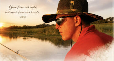 A serene lakeside scene at sunset features a person in a camo hat and sunglasses fishing. Text reads, "Gone from our sight, but never from our hearts."