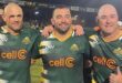 Three men in rugby jerseys stand smiling on a field. They link arms, showing camaraderie and joy. The stadium lights shine brightly in the background.